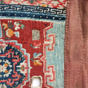 Textured red, blue, and cream patterned rug corner showing wear, stitched to a plain reddish-brown textile.