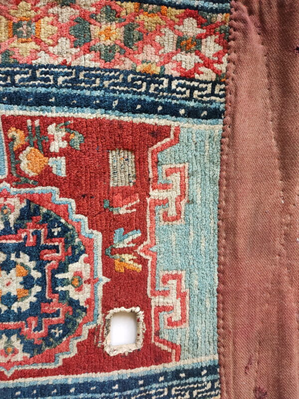 Textured red, blue, and cream patterned rug corner showing wear, stitched to a plain reddish-brown textile.