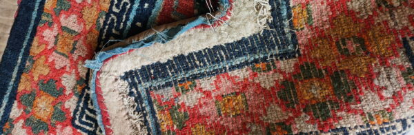Folded section of a textured, low-pile rug showing red, navy, green, and yellow patterns with exposed backing.