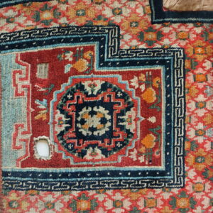 Highly patterned rug fragment featuring a red field, navy meander borders, gold flowers, and evident wear.