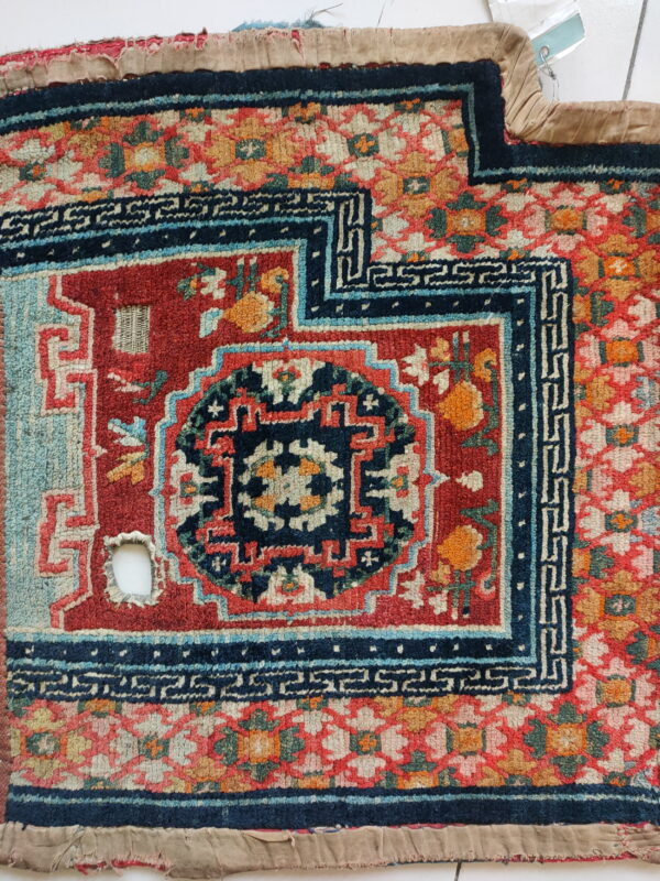 Highly patterned rug fragment featuring a red field, navy meander borders, gold flowers, and evident wear.