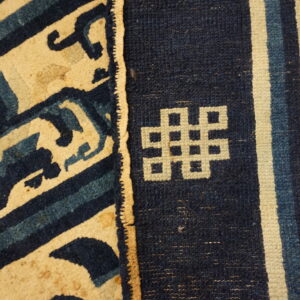 Low-pile rug detail showing indigo and cream geometric scrollwork separated from a knotted symbol and vertical stripes.