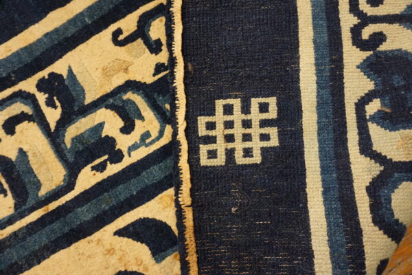 Low-pile rug detail showing indigo and cream geometric scrollwork separated from a knotted symbol and vertical stripes.