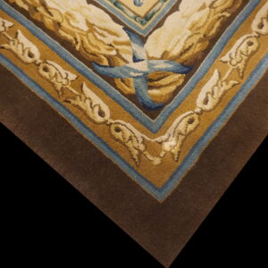 Close-up corner of a brown rug featuring tan scrolling borders and a central motif tied with a blue ribbon.