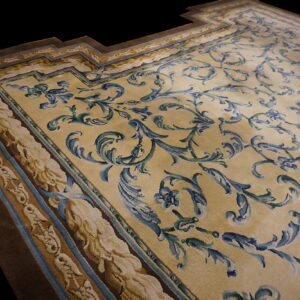 Pale gold rug with blue and green ornate scrolling designs, detailed borders, and a visible stepped corner.