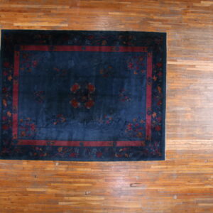 Dark blue low-pile rug with a red floral border and central medallion rests on a light brown plank wood floor.