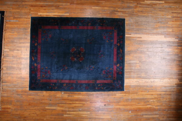 Dark blue low-pile rug with a red floral border and central medallion rests on a light brown plank wood floor.