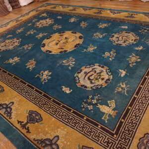 Turquoise and gold patterned rug with medallions rests on wood floor near stacks of rolled carpets.