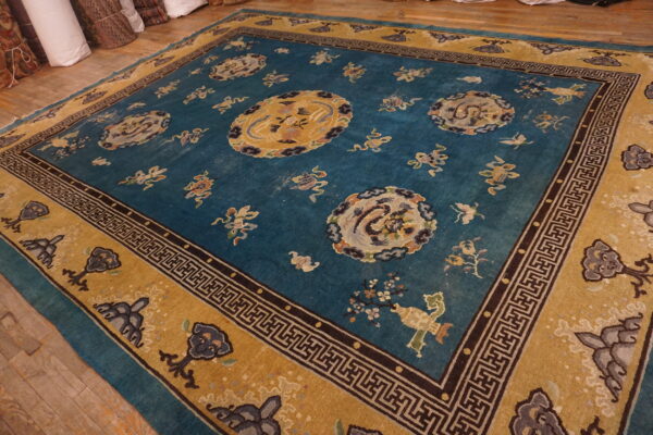 Turquoise and gold patterned rug with medallions rests on wood floor near stacks of rolled carpets.