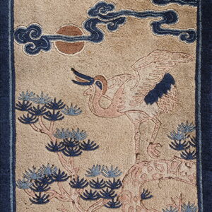 Tan rug with a dark blue border, patterned with a pink crane, blue pine needles, and stylized clouds.