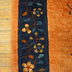 Orange rug edge with a navy floral border on distressed light wood plank flooring.