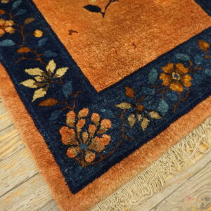 The burnt orange rug field meets a navy blue border with gold and teal floral motifs on wood planks.