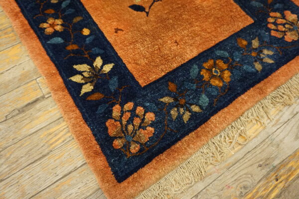 The burnt orange rug field meets a navy blue border with gold and teal floral motifs on wood planks.