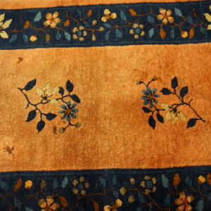 Rust-orange rug with a dark blue border featuring detailed multicolor floral and vine patterns.
