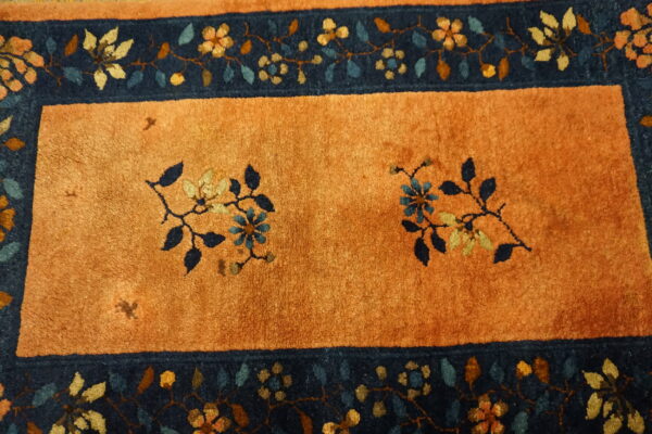 Rust-orange rug with a dark blue border featuring detailed multicolor floral and vine patterns.