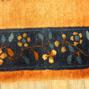 Orange rug detail showing a navy border patterned with gold flowers and blue leaves on wood planks.