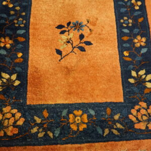 Rust-colored rug with a dark blue border featuring multicolored floral designs on distressed wood flooring.