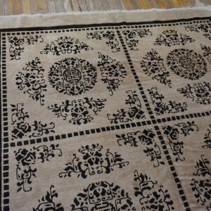 Taupe rug featuring dense black geometric and stylized floral motifs lies on weathered wood flooring.