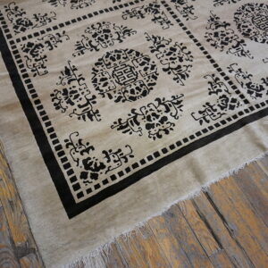 A light beige rug with a bold black geometric pattern and fringe lies on rustic wood plank flooring.