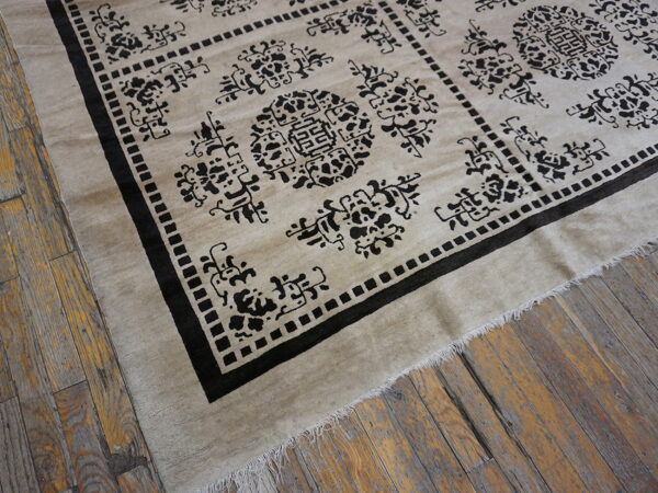 A light beige rug with a bold black geometric pattern and fringe lies on rustic wood plank flooring.