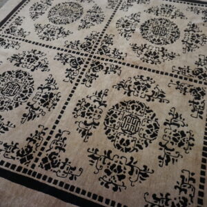 Tan rug featuring a repeating black geometric and stylized floral medallion pattern on distressed wood flooring.
