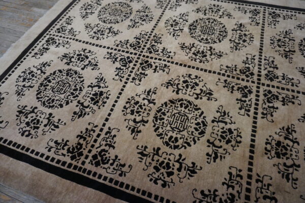 Tan rug featuring a repeating black geometric and stylized floral medallion pattern on distressed wood flooring.