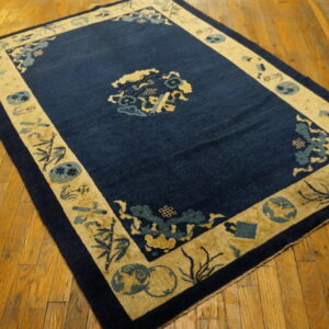 A rectangular rug featuring a dark blue field and light gold pictorial border is displayed on wood planks.