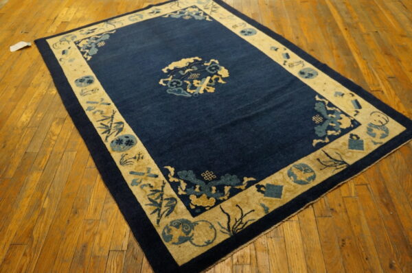 A rectangular rug featuring a dark blue field and light gold pictorial border is displayed on wood planks.
