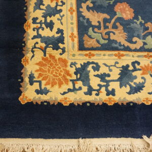 Navy blue rug corner showing cream and rust floral borders and white fringe on wood flooring.