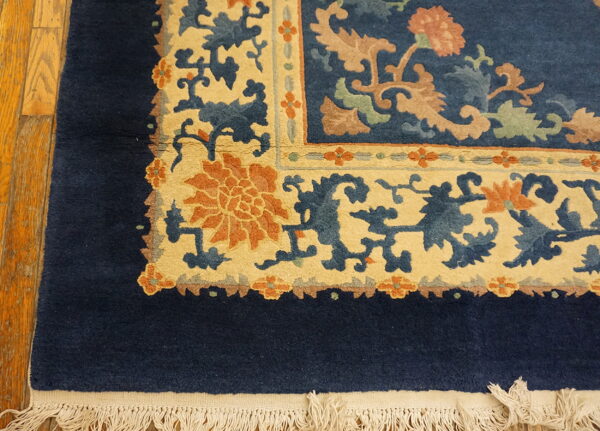 Navy blue rug corner showing cream and rust floral borders and white fringe on wood flooring.