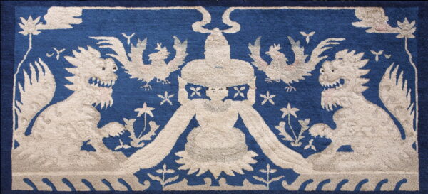 Low-pile blue rug with a beige design featuring two snarling beasts flanking a central figure.