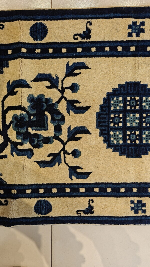 Short-pile tan rug with bold dark blue floral and geometric patterns rests on light square tiles.