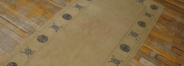 Low-pile tan rug featuring a repeating blue border design sits on worn, distressed hardwood planks.