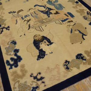 Beige rug with a dark blue border featuring a narrative pattern of figures around a table, lying on wood floorboards.
