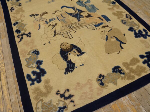 Beige rug with a dark blue border featuring a narrative pattern of figures around a table, lying on wood floorboards.