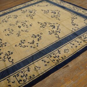 Tan area rug with scattered blue floral motifs and a Greek key border on rustic wood flooring.