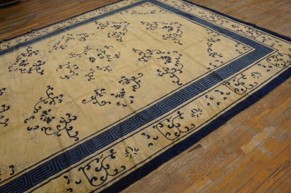 Tan area rug with scattered blue floral motifs and a greek key border on rustic wood flooring.