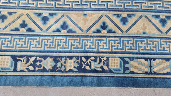 Detailed blue and beige rug borders show greek key and stylized floral patterns on a gray concrete floor.