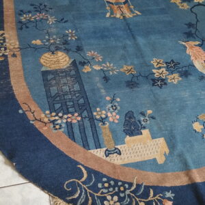 Oval low-pile blue rug with tan and navy borders features an ornate scenic and floral pictorial pattern on white tile.