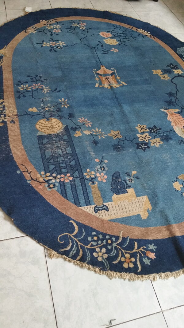 Oval low-pile blue rug with tan and navy borders features an ornate scenic and floral pictorial pattern on white tile.