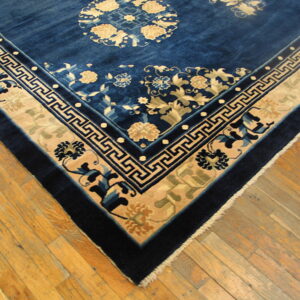 A dark blue rug with cream floral and Greek key borders rests on light-toned plank wood flooring.