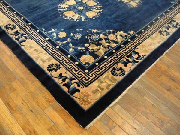 A dark blue rug with cream floral and greek key borders rests on light-toned plank wood flooring.