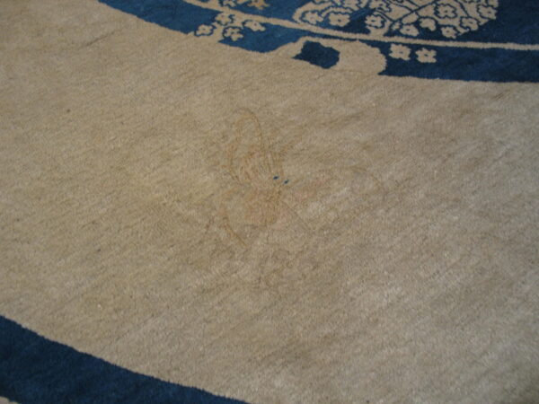 Close-up of a beige rug field with a faint reddish mark and a curved border of dark blue patterns.