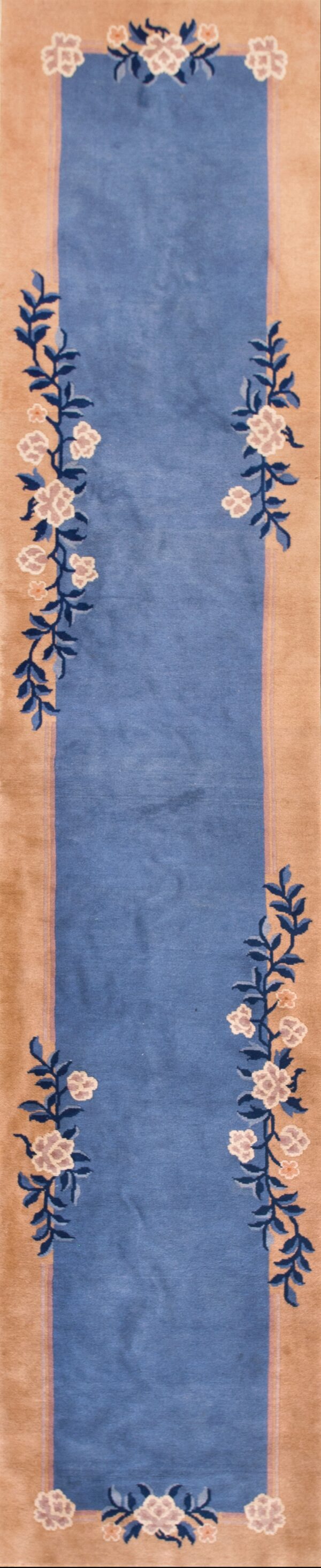 A rectangular blue rug features wide tan borders with floral and vine patterns, displayed on white tile.