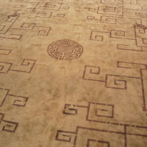Tan rug with dark brown key pattern repeating motifs and a circular labyrinth-like medallion.
