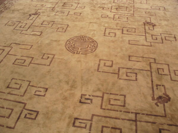 Tan rug with dark brown key pattern repeating motifs and a circular labyrinth-like medallion.
