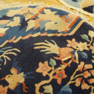 Dark blue, round rug with intricate peach, tan, and teal floral and bird motifs on distressed wood.