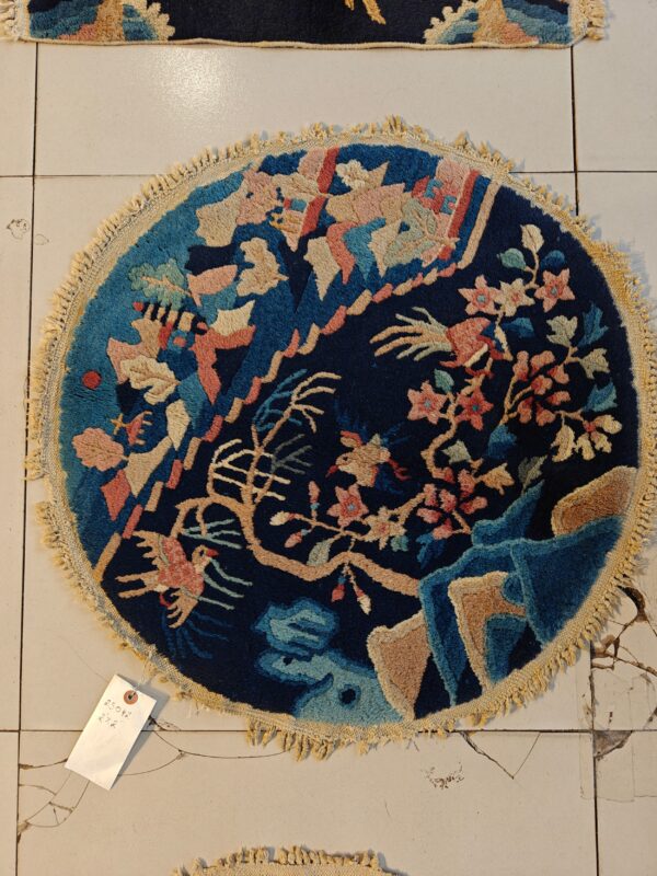 A circular navy rug with a high-pile floral and bird pattern in pinks, tan, and blues rests on cracked white tile.