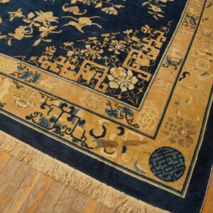 Corner of a dark blue and gold patterned rug with fringes, laid over aged hardwood floor planks.