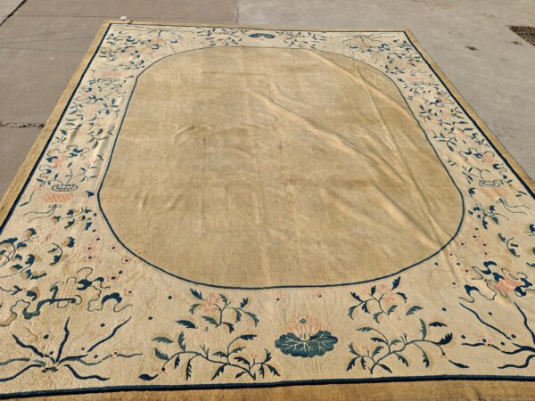 Muted tan rug with an oval field and blue and rose floral border laid out on gray pavement.